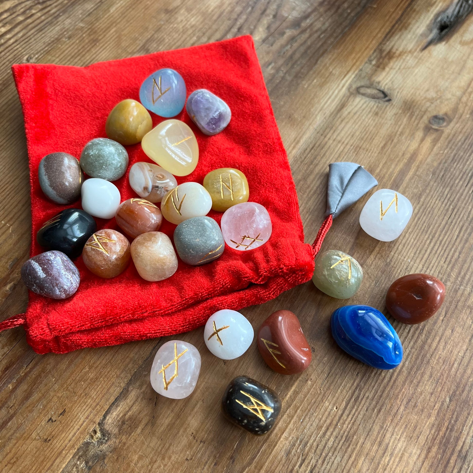 A set of polished tumbled gemstones with gold engraved Rune symbols, displayed on a red velvet pouch and scattered on a wooden tabletop.