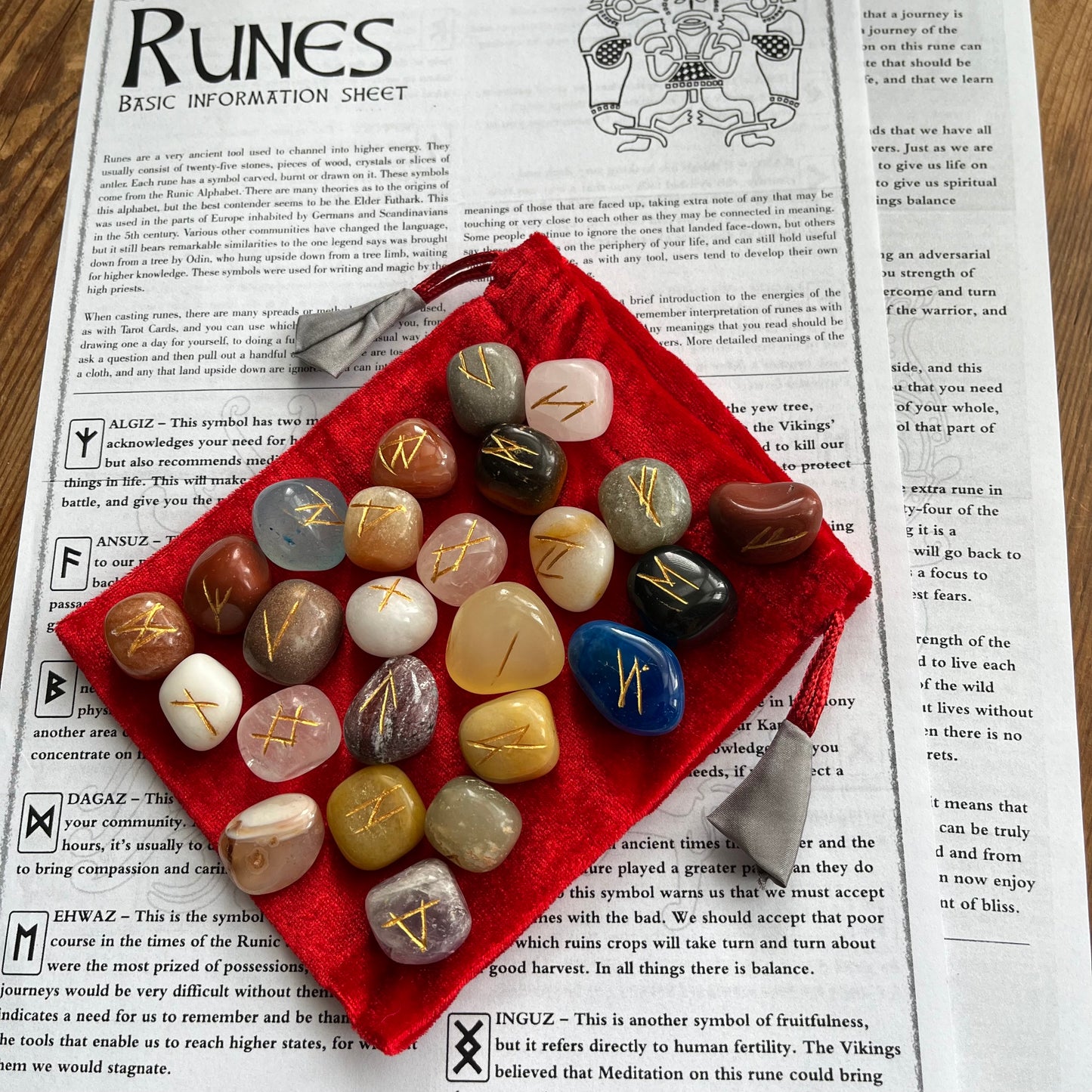 A set of 25 polished gemstone runes with engraved gold symbols resting on a red velvet bag placed on top of an open book showing pages about the Runes.