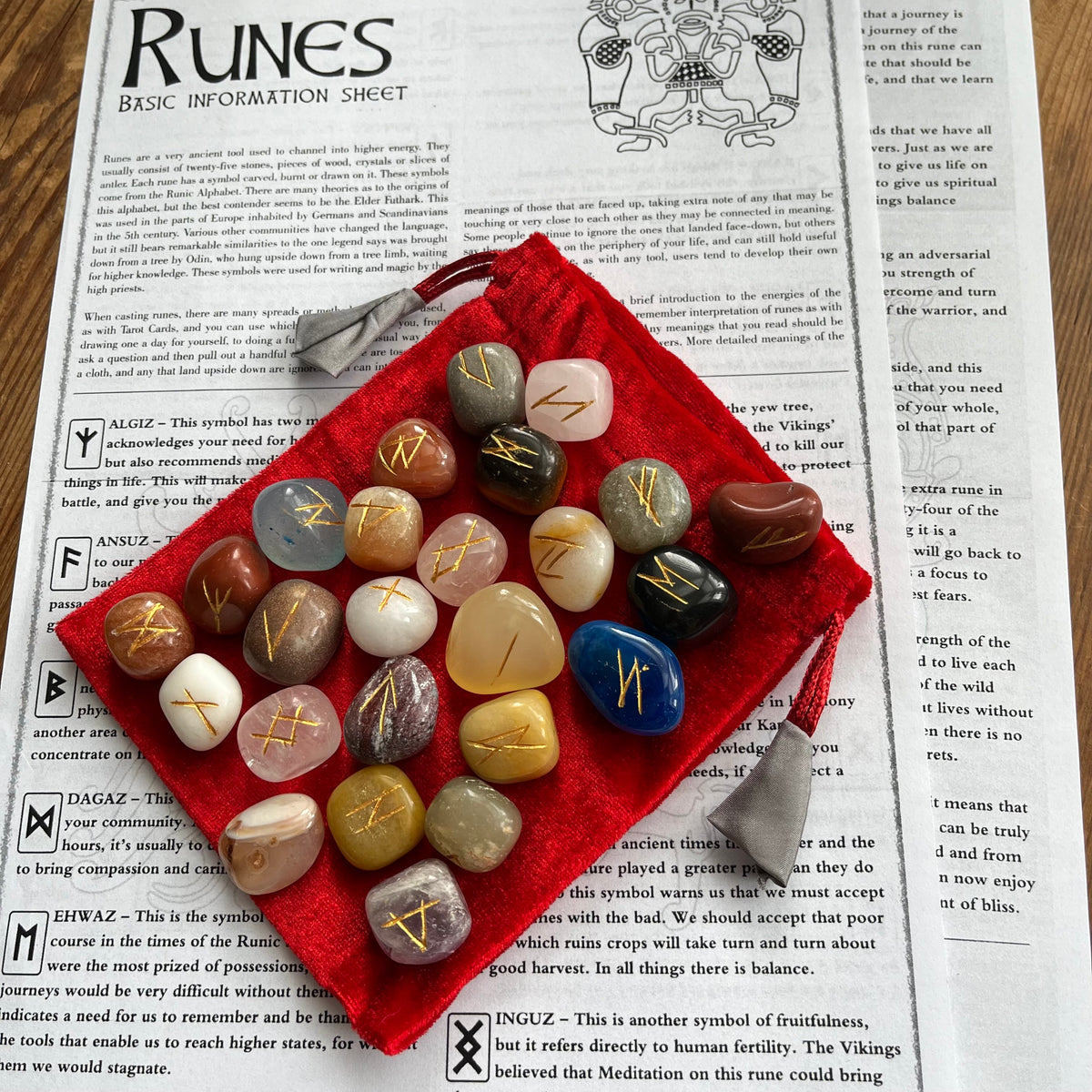 A set of 25 polished gemstone runes with engraved gold symbols resting on a red velvet bag placed on top of an open book showing pages about the Runes.
