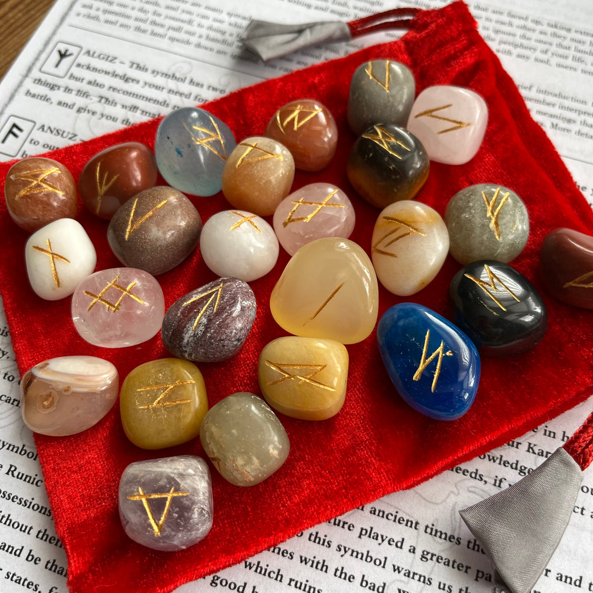 A set of polished tumbled gemstones with engraved gold Rune symbols, arranged on a red velvet drawstring pouch over printed book pages.