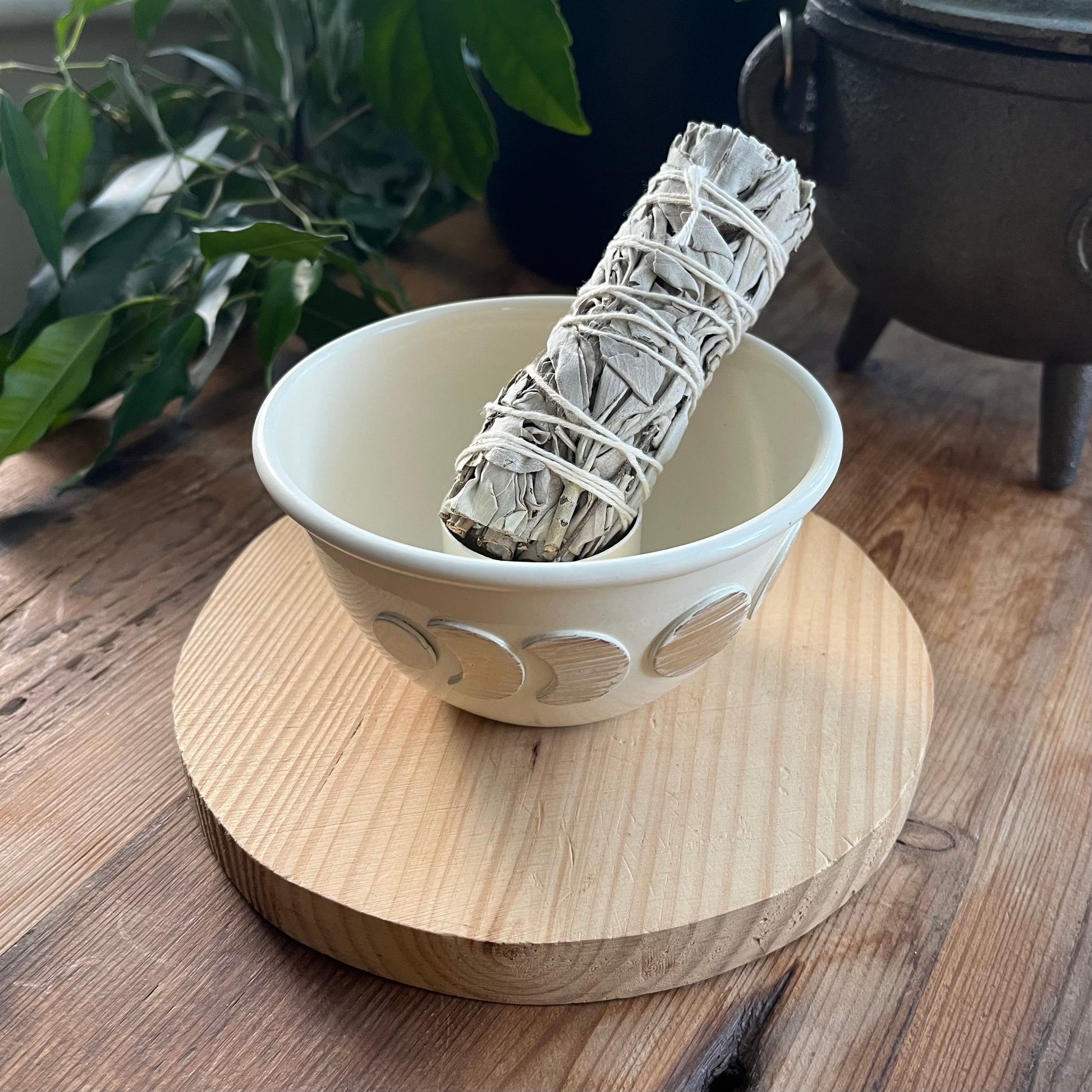 White ceramic bowl with a sage smudge stick on a wooden surface, with plants and a cauldron in the background.