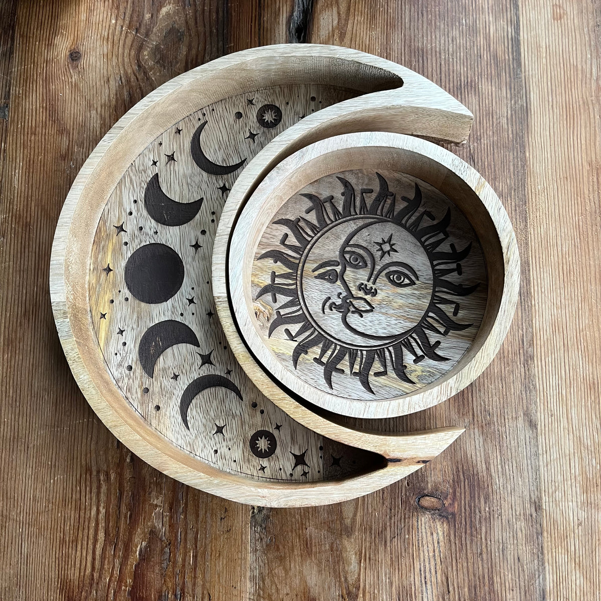 A set of two nested handcrafted wooden bowls with celestial designs, including a large crescent moon, a medium bowl featuring a detailed sun and moon face, displayed on a rustic wooden surface.