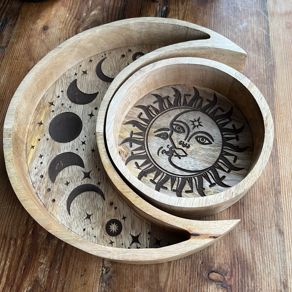 Set of two wood trays. The larger crescent moon tray features engraved moon phases and contains crystals. A smaller round tray, decorated with a radiant sun and moon motif contains a tarot deck. Both are nestled together and arranged on a rustic wooden table next to a potted succulent.