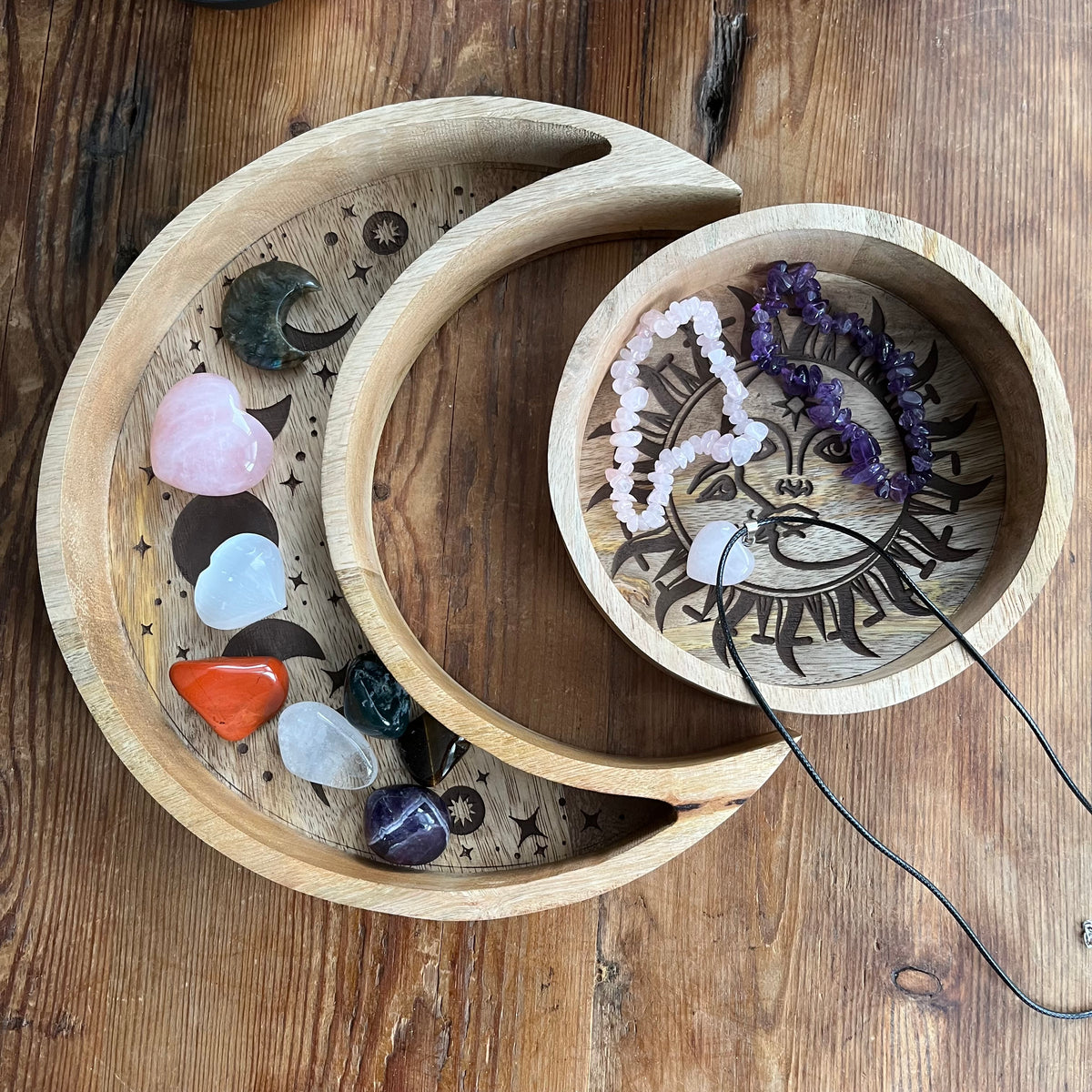 Set of two wood trays. The larger crescent moon tray features engraved moon phases and contains crystals. A smaller round tray, decorated with a radiant sun and moon motif contains crystal jewellery. Both are nestled together on a wooden table.
