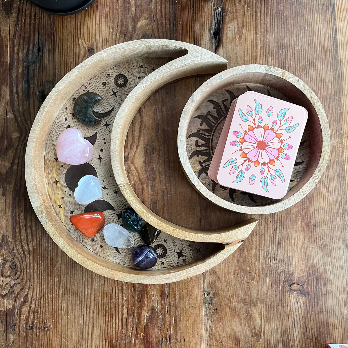 Set of two wood trays. The larger crescent moon tray features engraved moon phases and contains crystals. A smaller round tray, decorated with a radiant sun and moon motif contains a tarot deck. Both are nestled together on a wooden table.