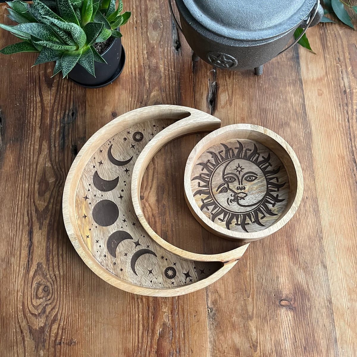 Set of two wood trays. The larger crescent moon tray features engraved moon phases and contains crystals. A smaller round tray, decorated with a radiant sun and moon motif contains a tarot deck. Both are nestled together and arranged on a rustic wooden table next to a potted succulent.