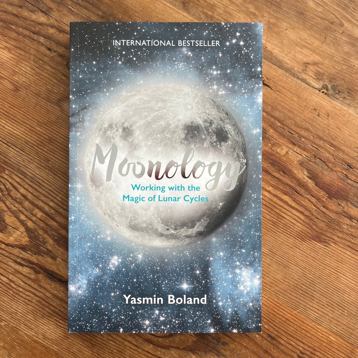 The Front cover of the book 'Moonology' by Yasmin Boland, displayed flat on a brown wooden surface. The cover features a dark blue, starry night sky background with glowing moon, white text, and a barcode at the bottom.