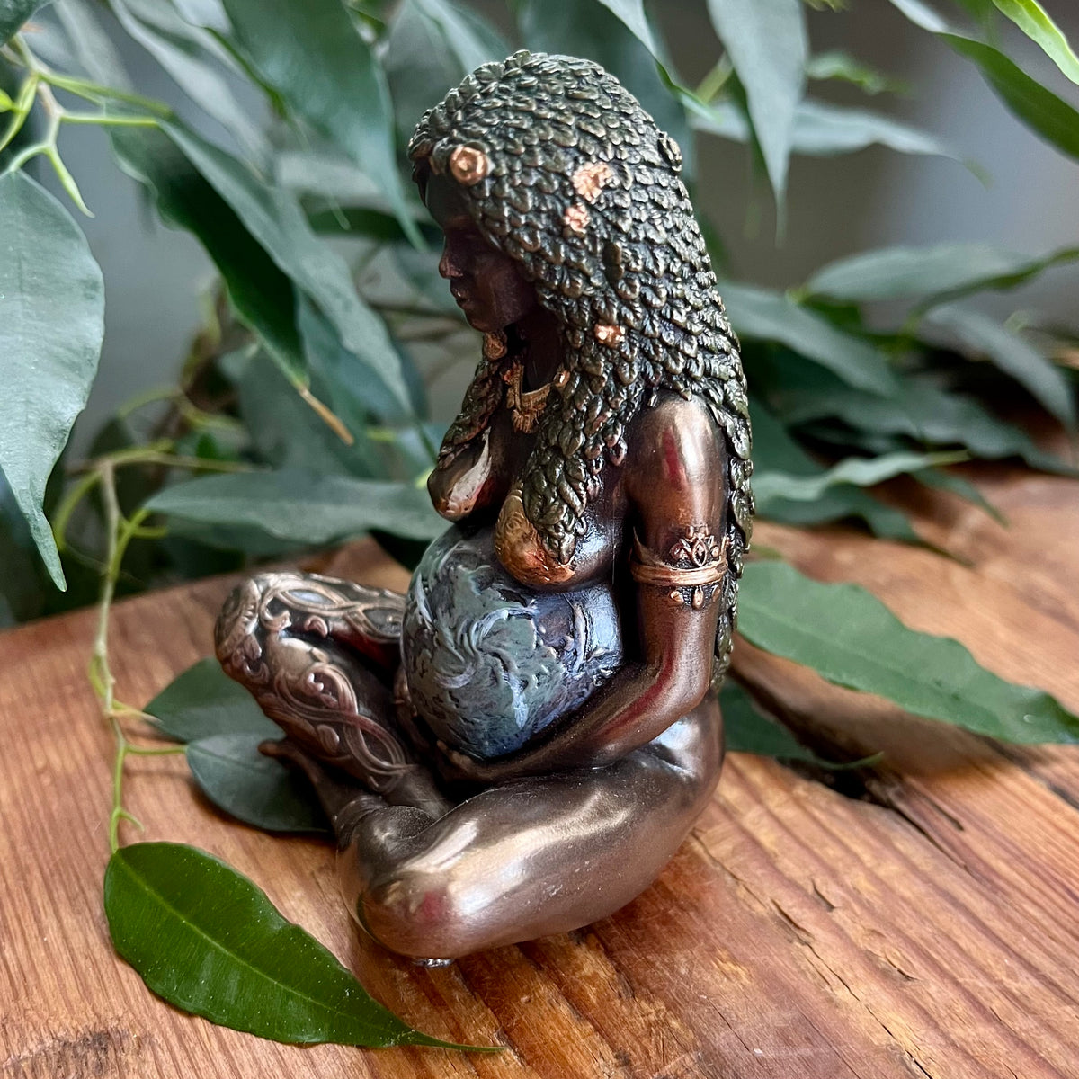 A small bronze figurine of a seated nude female figure with textured hair and jewelry, holding a blue-gray globe, displayed on a wooden surface with green leaves in the background.