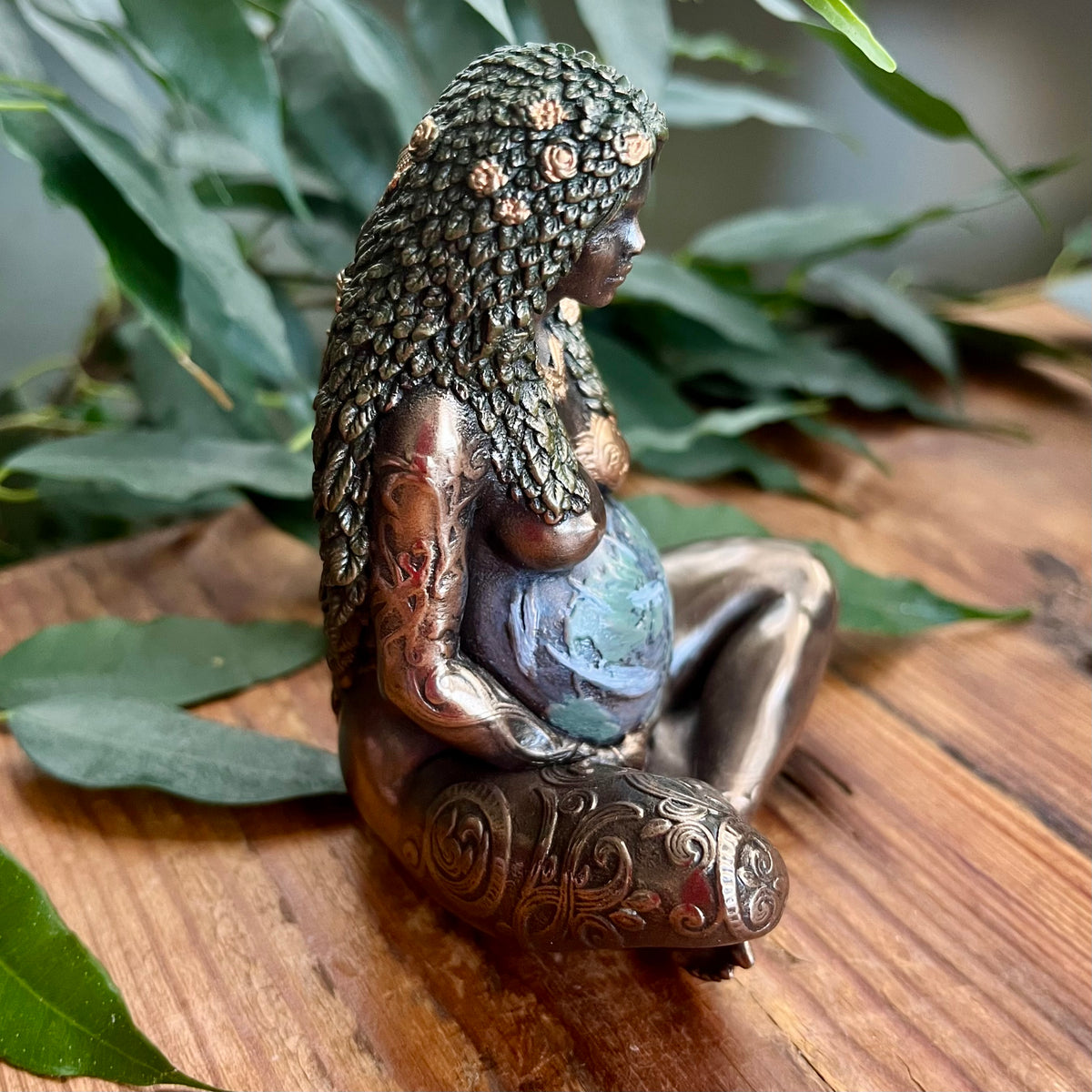 A detailed figurine of a pregnant goddess with long textured hair adorned with flowers, sitting cross-legged on a wooden surface with green leaves in the background.
