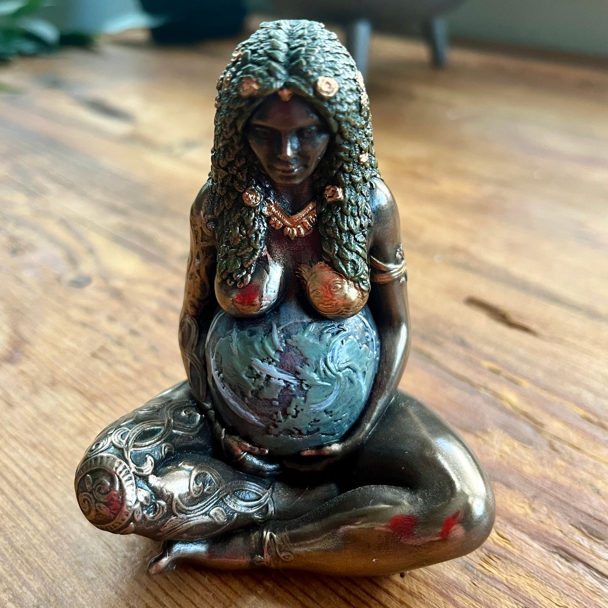 A bronze-finished figurine of a seated pregnant woman holding a globe, displayed on a wooden tabletop.