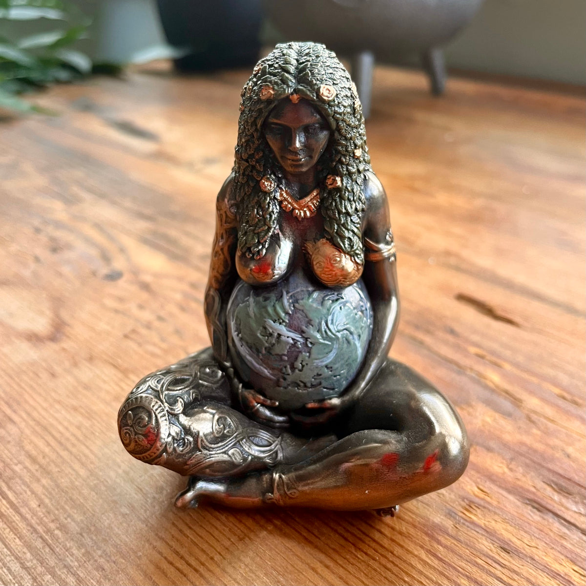 A bronze-toned figurine of a pregnant goddess sitting cross-legged and holding a globe, displayed on a wooden tabletop.