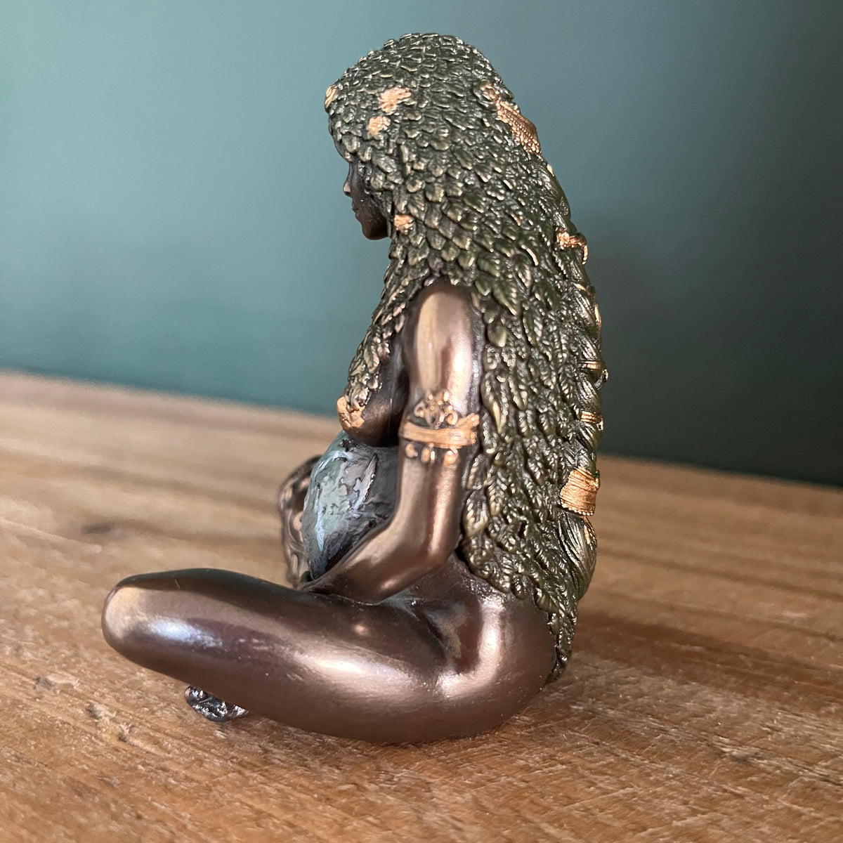 A bronze-finished figurine of a seated pregnant woman with textured hair, shown from the side on a wooden surface against a dark teal background.