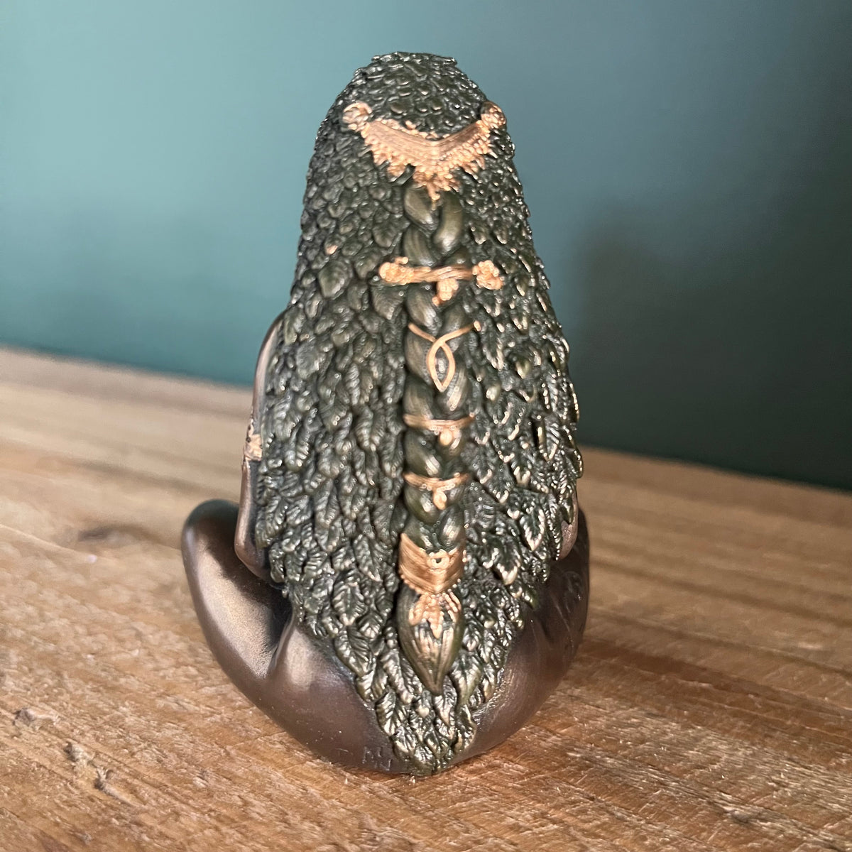 A detailed fantasy figurine of a seated figure viewed from behind, featuring long dark textured hair with a braided center and metallic bronze accents, displayed on a wooden surface against a dark teal background.