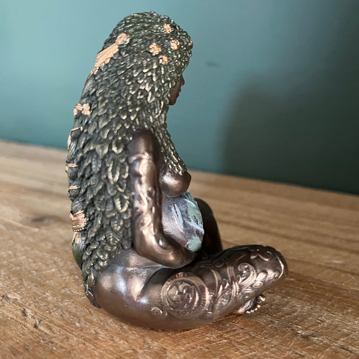 A small bronze figurine of a seated female figure with textured hair photographed on a wooden surface against a teal wall.