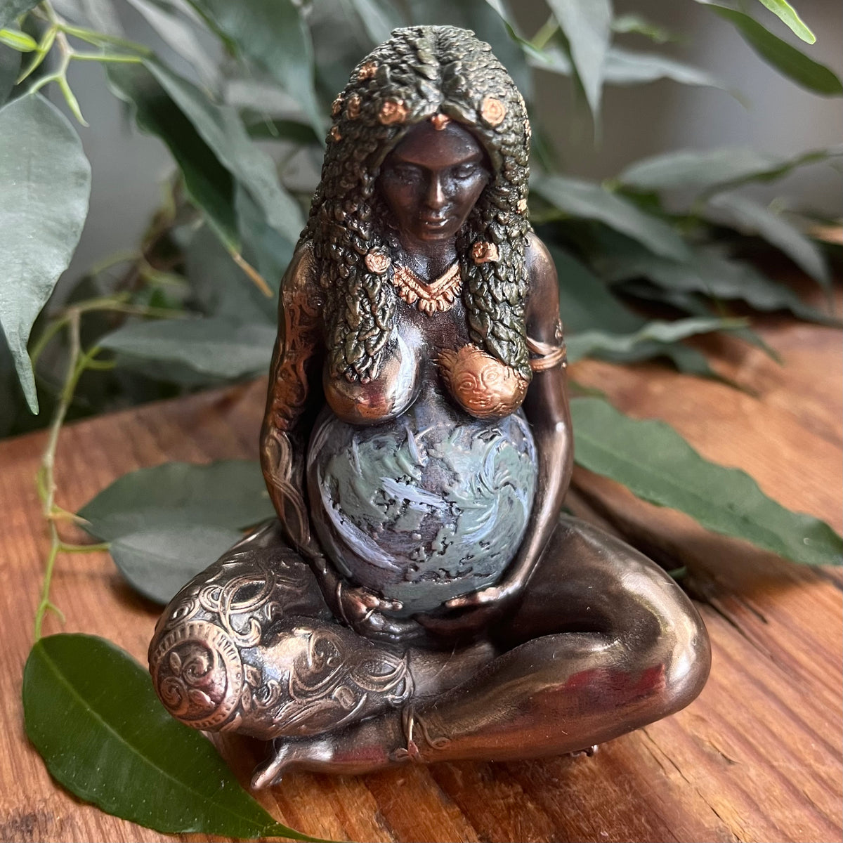 A bronze-finished figurine of a pregnant goddess sitting cross-legged and cradling a baby globe, styled on a wooden surface with green leaves in the background.