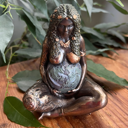 A bronze-finished figurine of a pregnant goddess sitting cross-legged and cradling a baby globe, styled on a wooden surface with green leaves in the background.