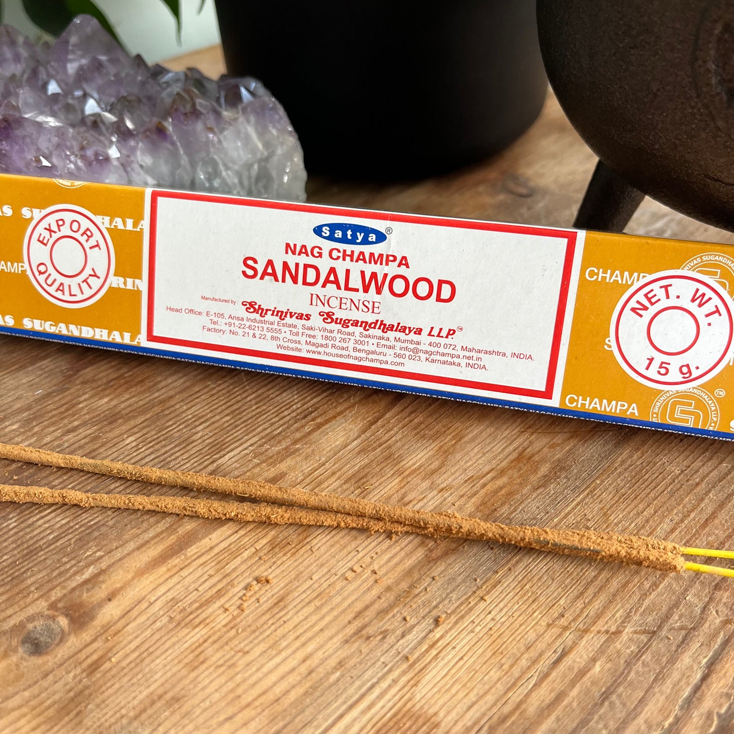 A box of Satya Nag Champa Sandalwood incense, with branding and product information displayed on the packaging, placed on a wooden surface. Alongside some loose incense sticks