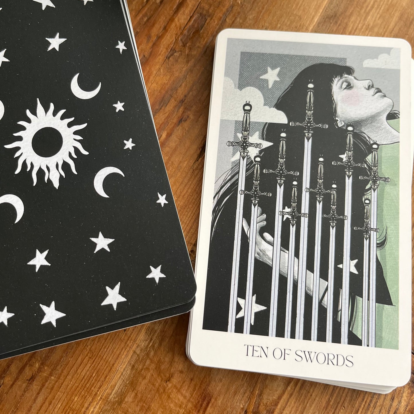 A tarot deck of cards on a wooden surface, showing the back of the card box with white celestial symbols on a black background, and the 'Ten of Swords' card featuring an illustration of a woman with swords piercing her chest.