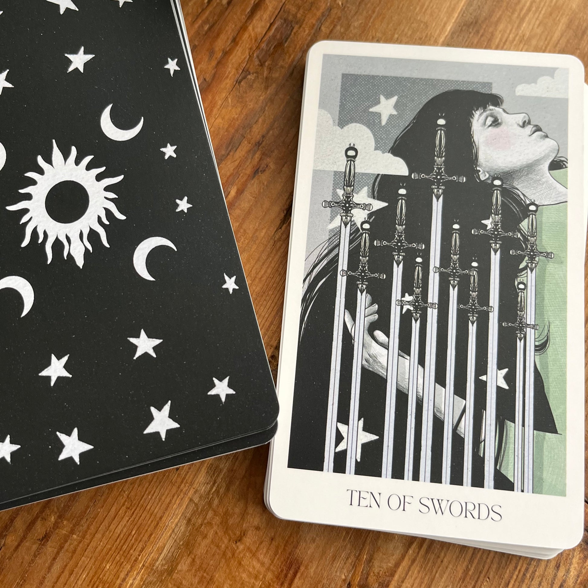 A tarot deck of cards on a wooden surface, showing the back of the card box with white celestial symbols on a black background, and the 'Ten of Swords' card featuring an illustration of a woman with swords piercing her chest.