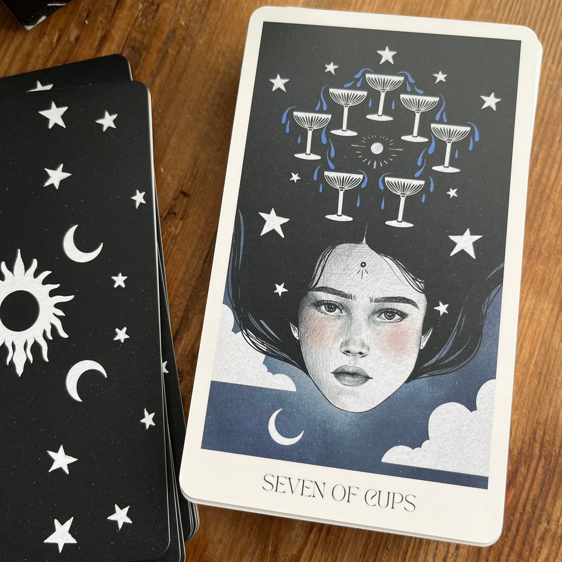 A tarot card deck on a wooden surface, showing the 'Seven of Cups' card with an illustration of a woman's face, stars, and cups, next to the back of another card with celestial designs.