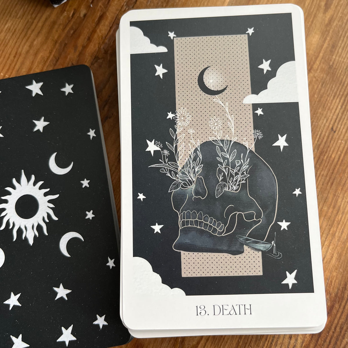 A tarot deck card labeled '13. DEATH' lies on a wooden surface beside another card showing celestial motifs like stars, moons, and a sun.