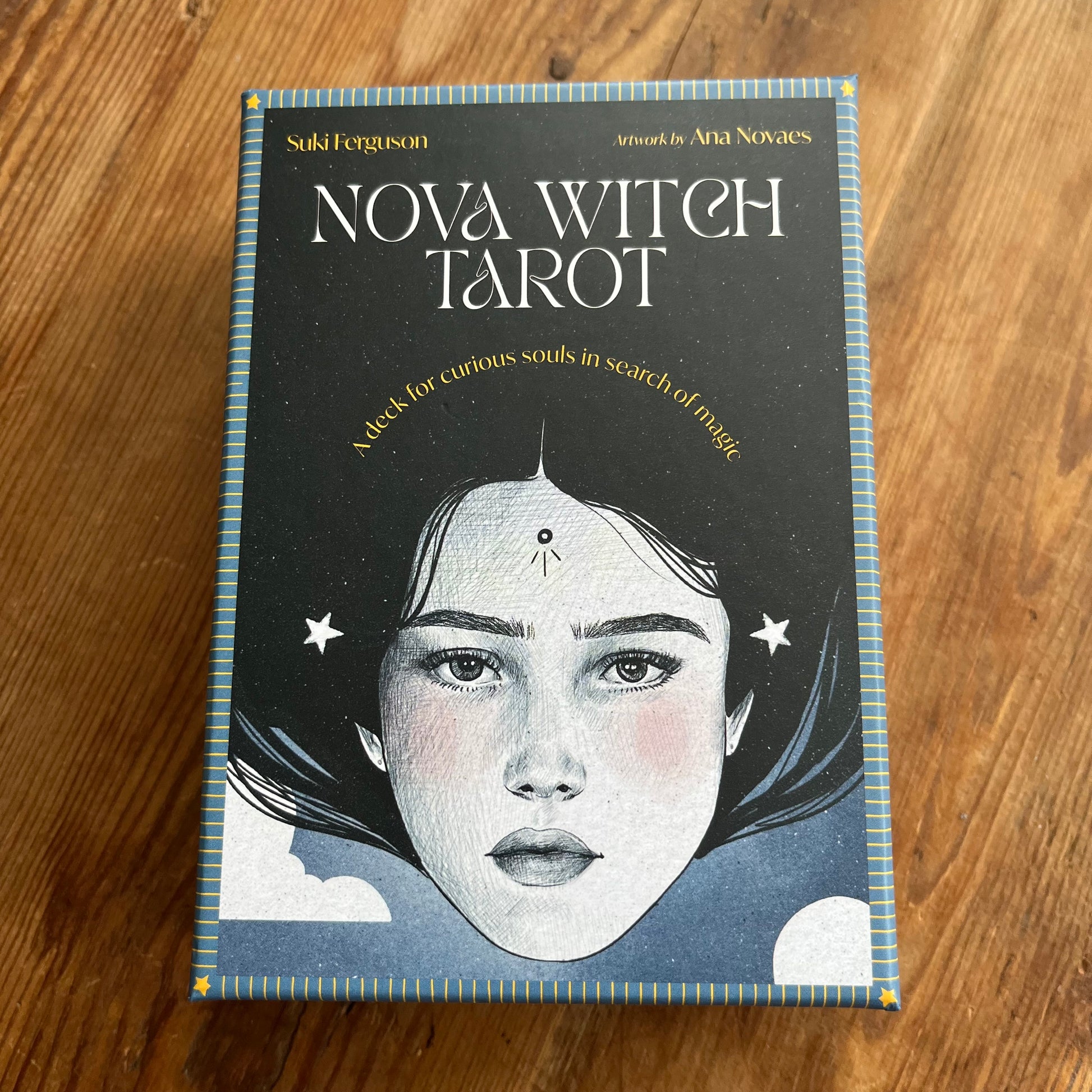 The 'Nova Witch Tarot' deck box is placed flat on a wooden table. The box features a dark, starry background with a stylized illustration of a woman's face, white text, and a blue border.