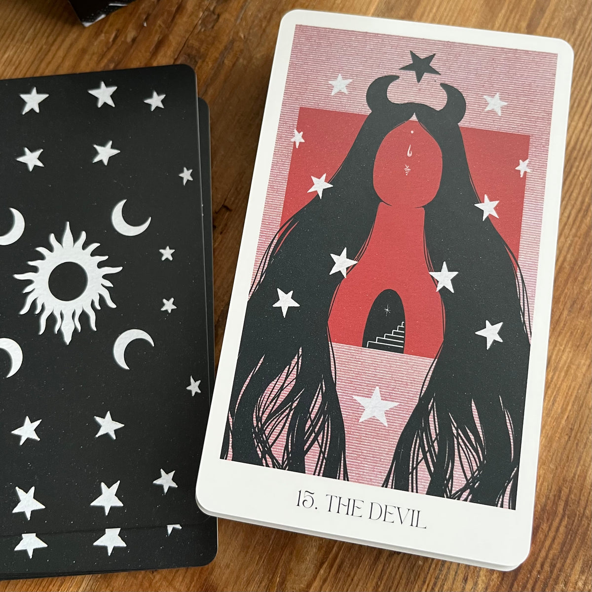 A tarot card deck on a wooden surface, showing the front of card 15 'The Devil' with a red and black illustration, and the back of another card featuring white celestial symbols on a black background.