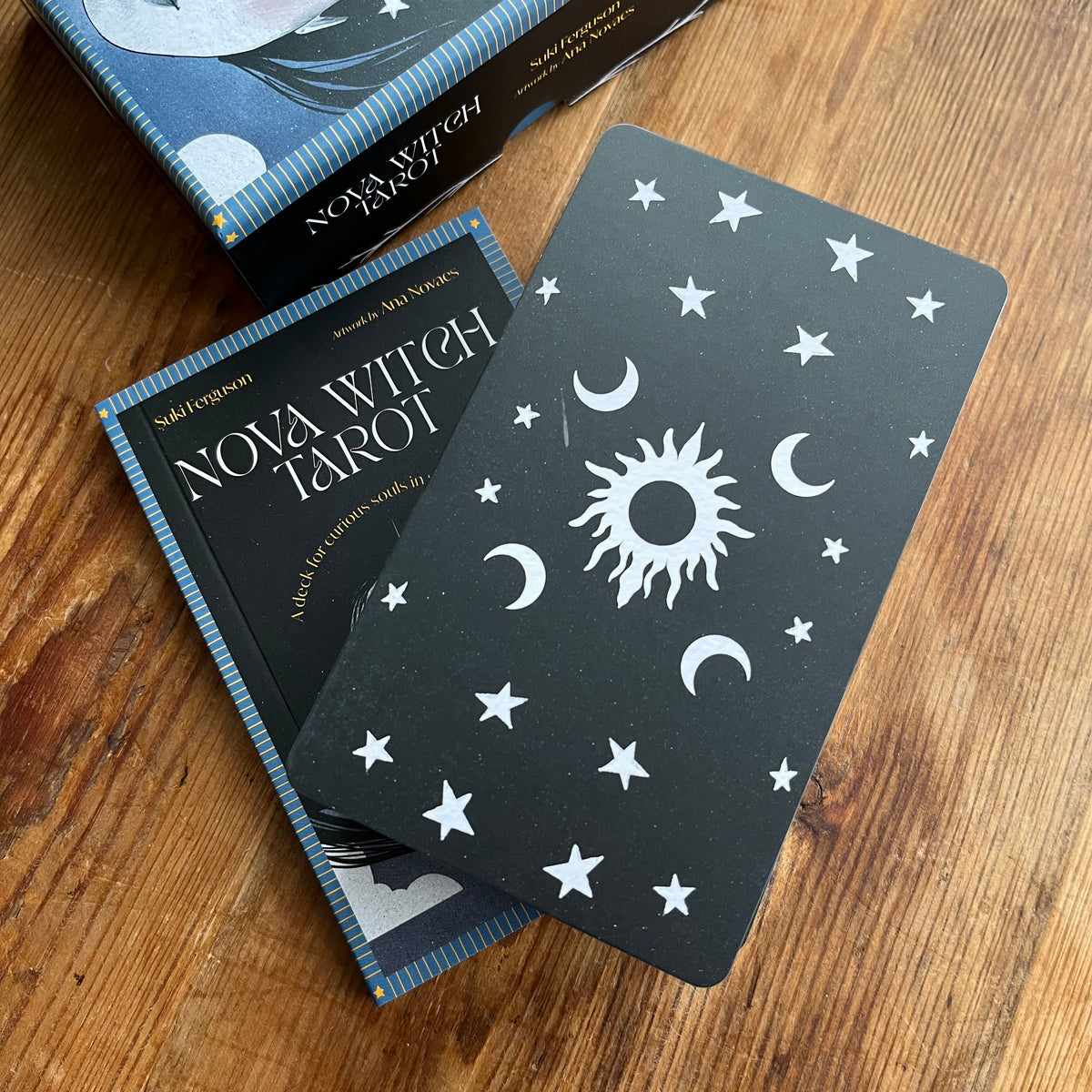 A Noxa Witch Tarot deck and card set displayed on a wooden tabletop, featuring a black card with white celestial illustrations and the blue-and-black box labeled 'Nova Witch Tarot'.