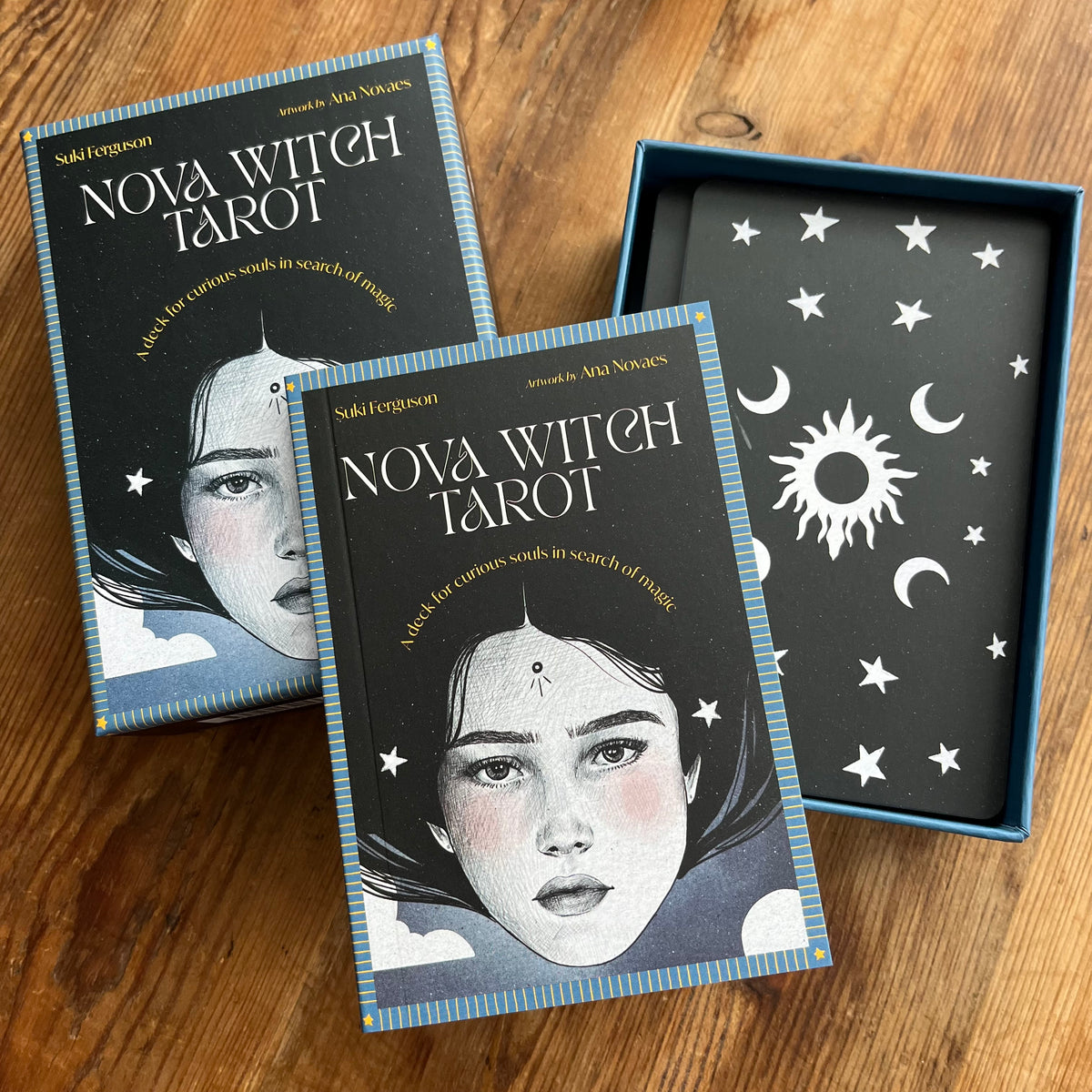 Nova Witch Tarot deck and its box displayed on a wooden surface, with two tarot cards in front and an open box showing the black card back decorated with white celestial symbols.
