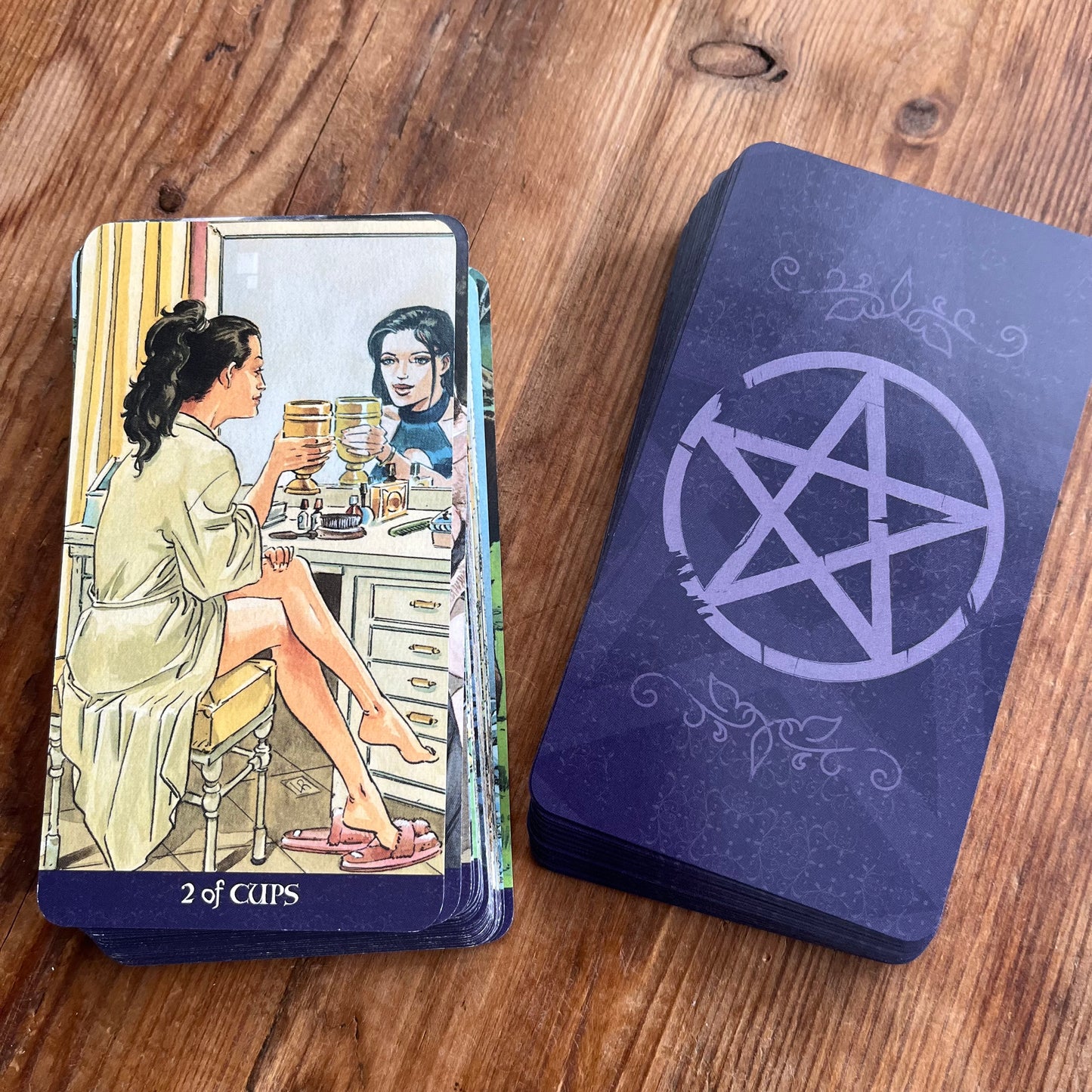 A tarot card deck with illustrated cards showing '2 of Cups' and a dark blue-purple back with a pentagram symbol, placed on a wooden tabletop.