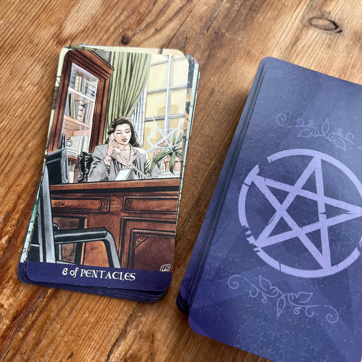A tarot card deck on a wooden tabletop, showing the '8 of Pentacles' card face and the blue-backed card stack with a white pentagram symbol.