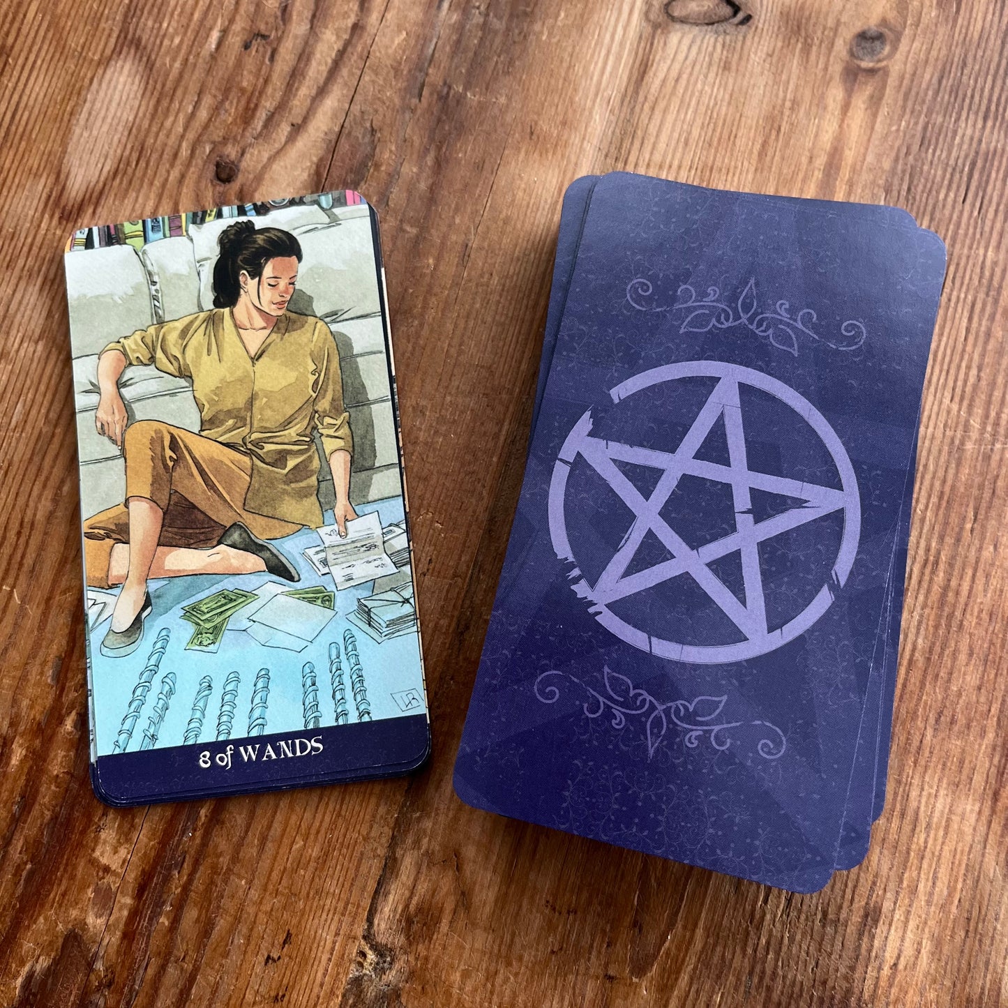 A tarot deck with the '8 of Wands' card face-up and a stack of cards with a dark blue background and a white pentagram design, placed on a wooden tabletop.