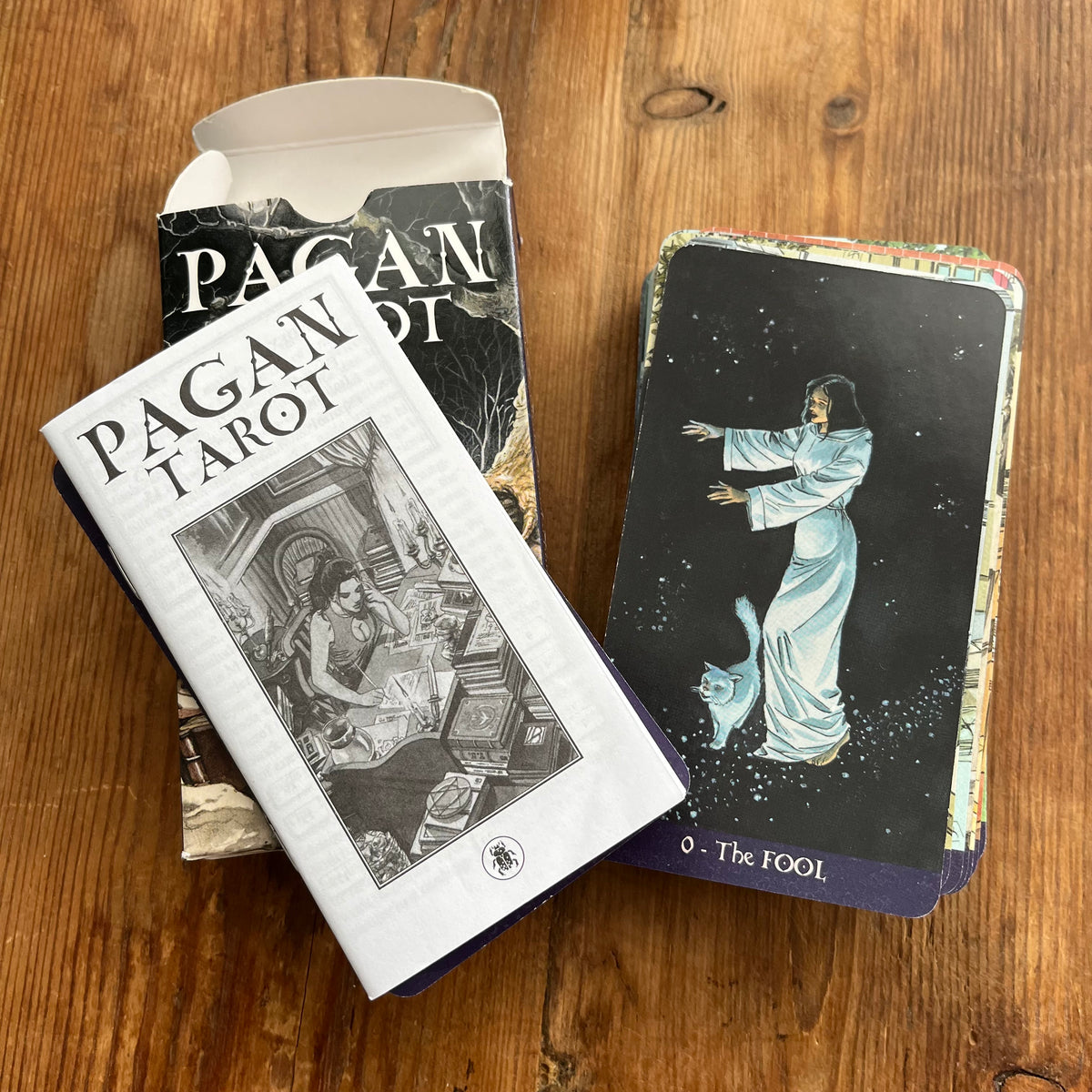 A Pagan Tarot deck displayed on a wooden table, showing the white cover with black text, the open black-and-white box, and a tarot card featuring a figure in a white robe on a dark background.