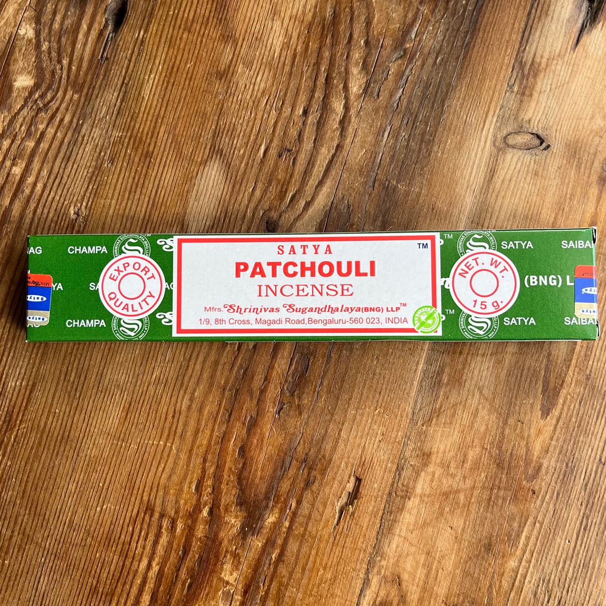 A rectangular box of Satya Patchouli Incense placed horizontally on a rustic wooden surface. The box is primarily green with a large white center panel featuring red text and circular red and green logos.