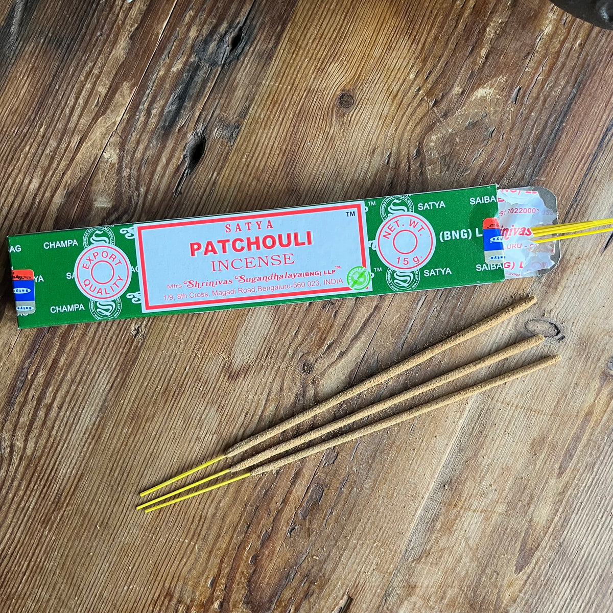 A package of Satya Patchouli Incense sticks lies on a rustic wooden tabletop, with several incense sticks placed in front of it.