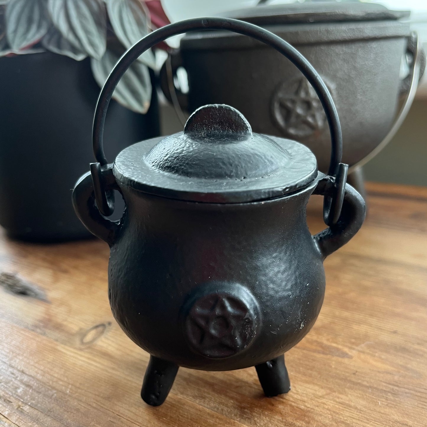 A small black cast-iron cauldron with a lid, loop handle, and triple feet, featuring an embossed pentagram symbol on the front, displayed on a wooden tabletop.