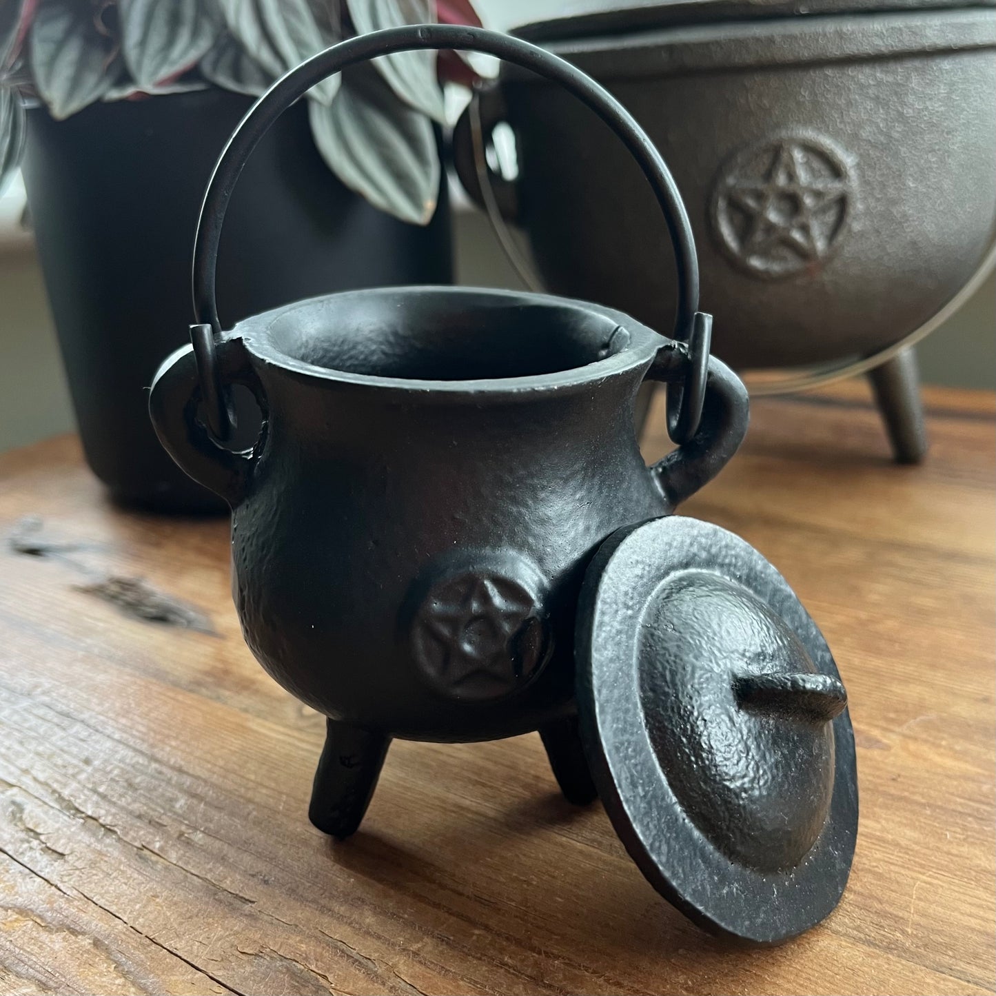 A black cast iron cauldron-shaped container with a lid leaning against it, placed on a wooden surface. The cauldron has a triple foot base, a top handle, and an embossed pentagram symbol on the side.