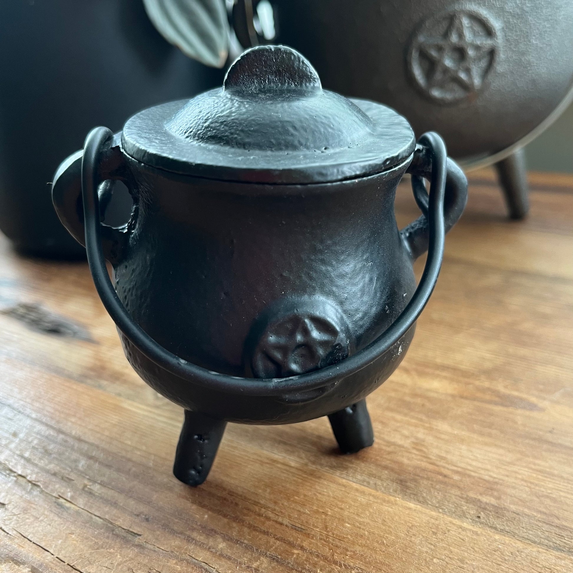 A small black cast iron cauldron-shaped pot with a lid, loop handles, and a triple foot base, sitting on a wooden tabletop with a larger dark object blurred in the background.