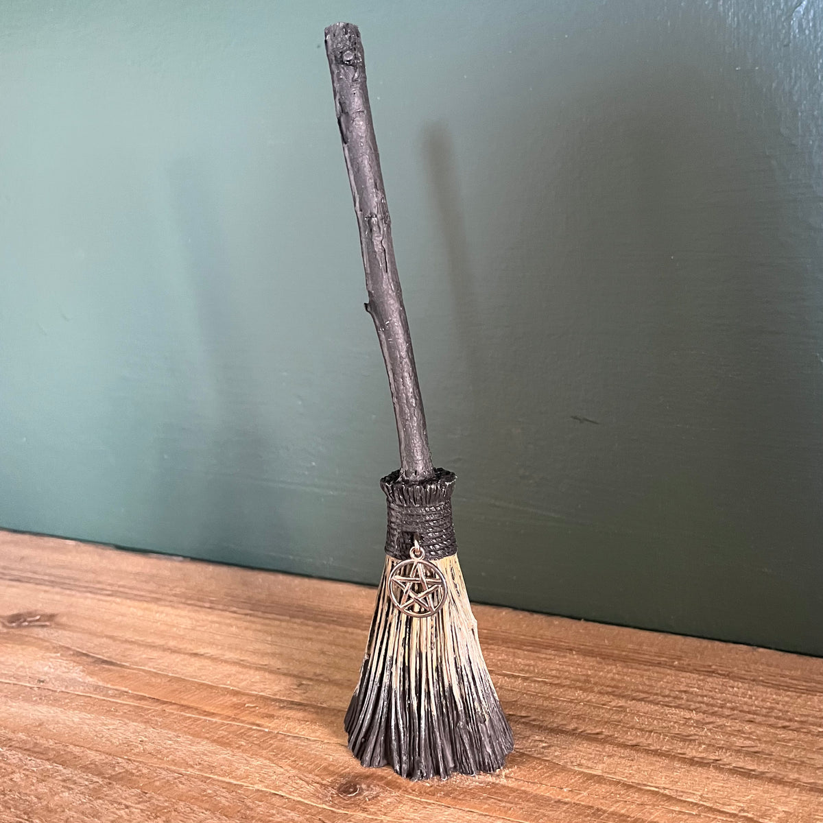 A miniature decorative broomstick figurine with a dark brown faux-wood handle, black bristles, and a silver pentagram ornament, standing on a wooden surface against a dark green wall.
