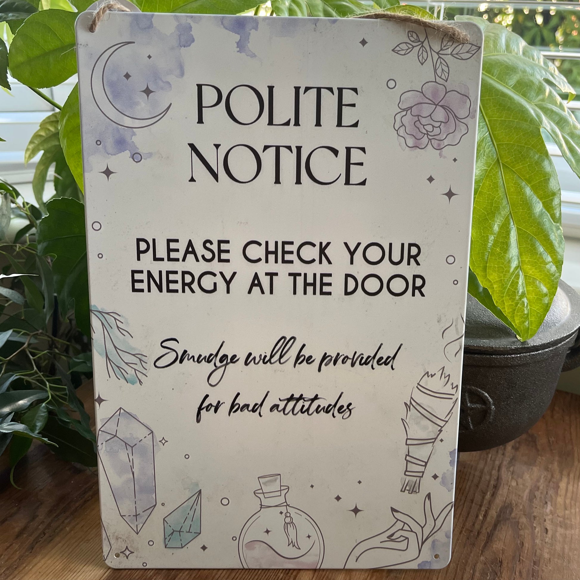 Decorative fun witchy sign with 'Polite Notice' to check your energy text and illustrations on a wooden surface with plants in the background.