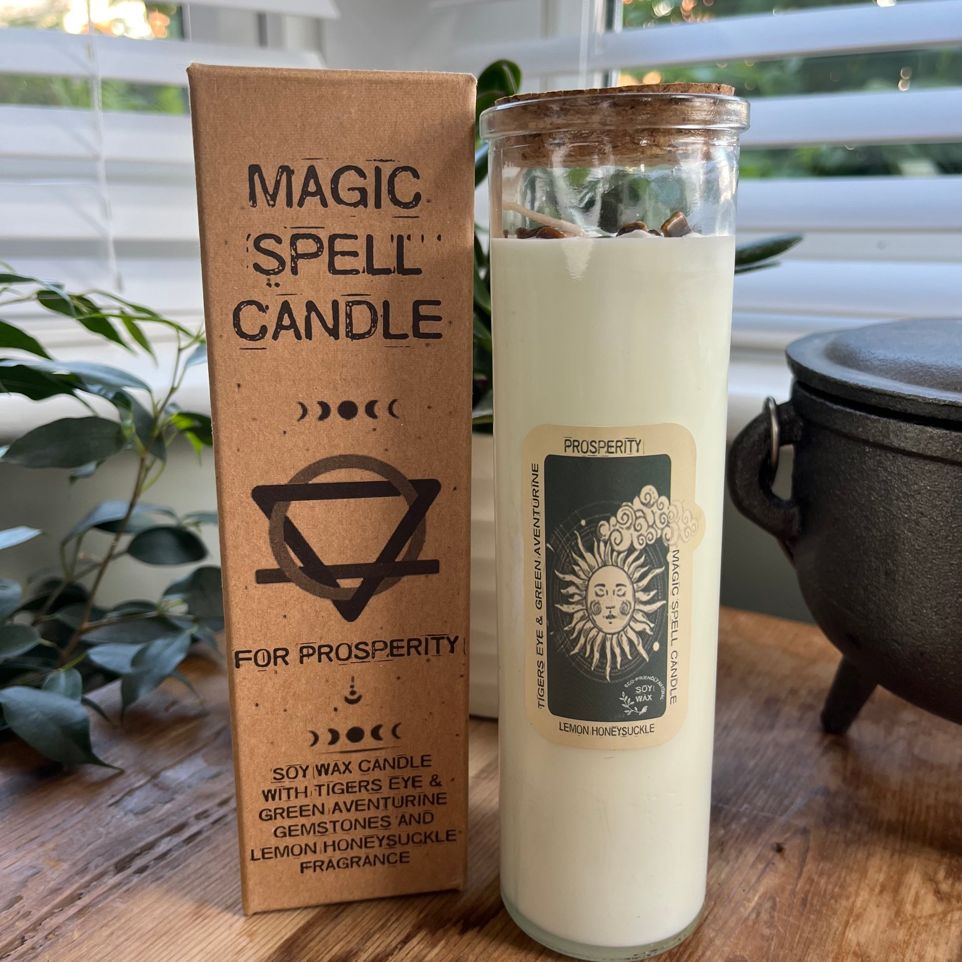 Tall glass spell candle with cork lid. The label on the candle features an image of the sun and the words Prosperity, Tigers Eye And Green Aventurine and Lemon Honeysuckle. The candle itself is White with Tigers Eye and Green Aventurine gemstones embedded within it. Alongside the glass Magic spell candle is it's brown cardboard box which has the words magic spell candle for Prosperity, soy wax candle with Tigers Eye and Green Aventurine gemstones and Lemon Honeysuckle fragrance.