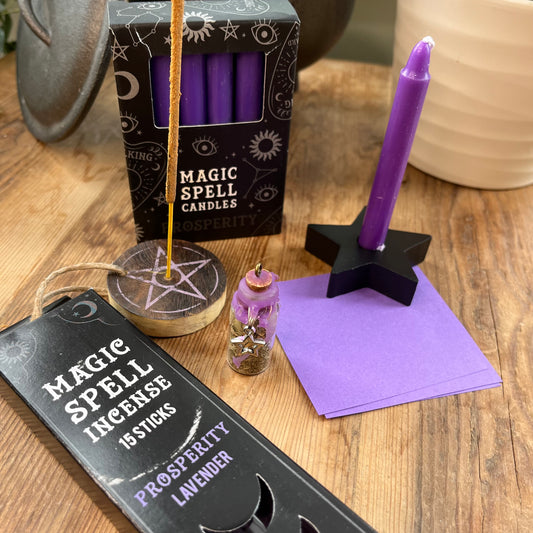 'Prosperity Spell Kit' Contents displayed on a wooden table. Pack of lavender scented incense sticks for prosperity, and wooden pentagram incense holder, Pack of purple magic spell candles, wooden black star shaped candle holder, squares of purple paper and a "prosperity' spell bottle containing herbs and sealed with purple wax and a charm.