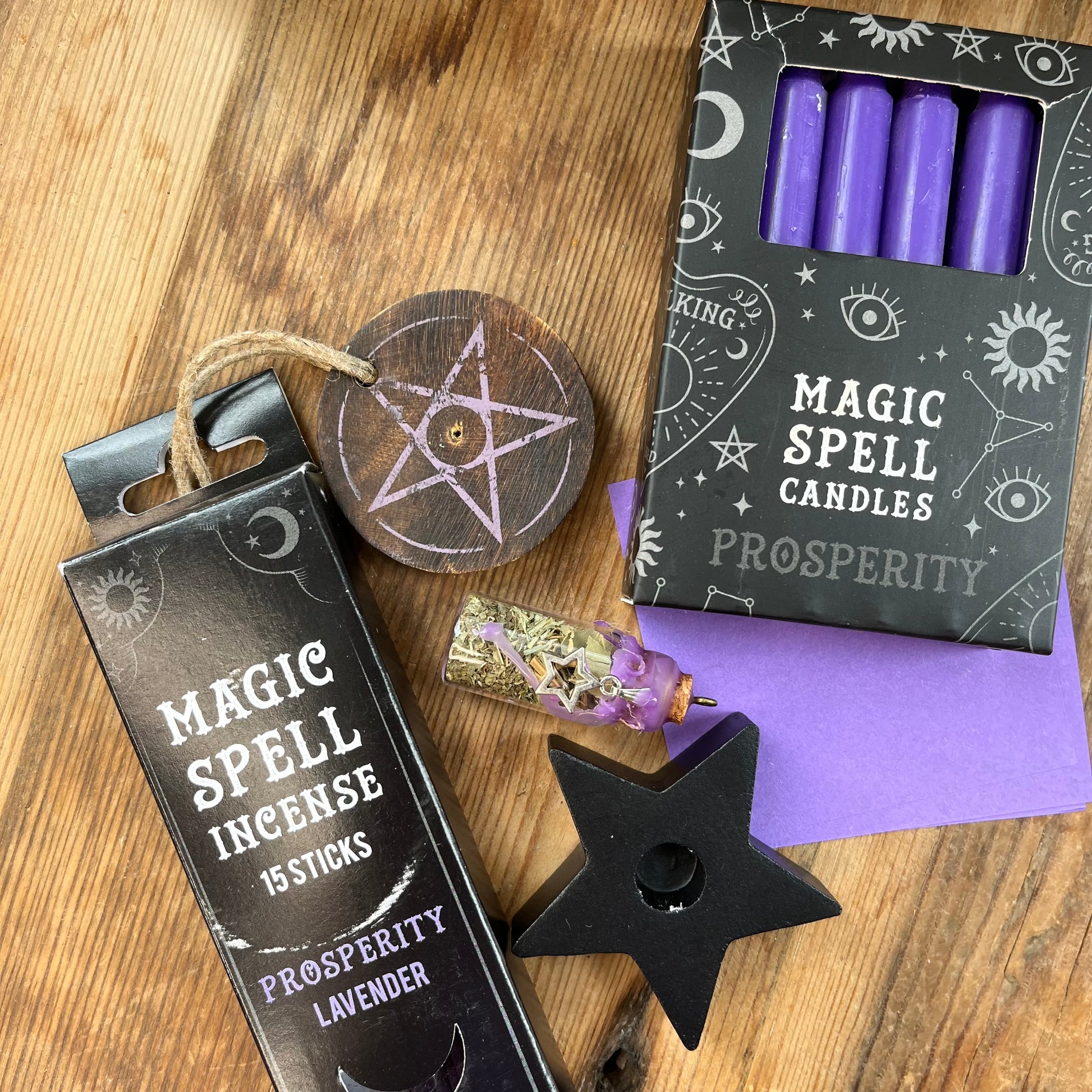 'Prosperity Spell Kit' Contents displayed on a wooden table. Pack of lavender scented incense sticks for prosperity, and wooden pentagram incense holder, Pack of purple magic spell candles, wooden black star shaped candle holder, squares of purple paper and a "prosperity' spell bottle containing herbs and sealed with purple wax and a charm.