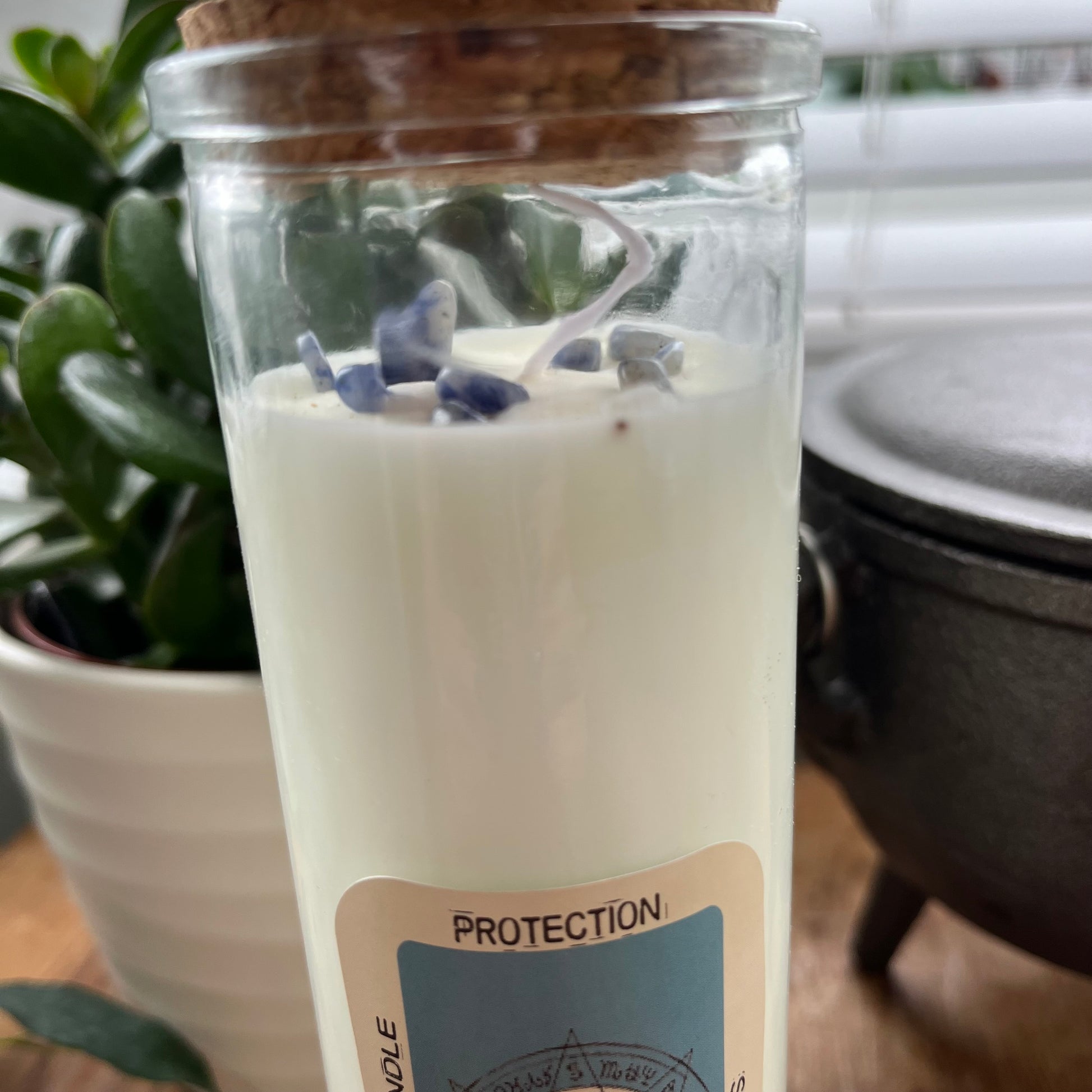 Tall glass spell candle with cork lid. The label on the candle features an image of the all seeing eye and the words Protection Sodalite and Dragons Blood. The candle itself is white with Sodalite gemstones embedded within it. 