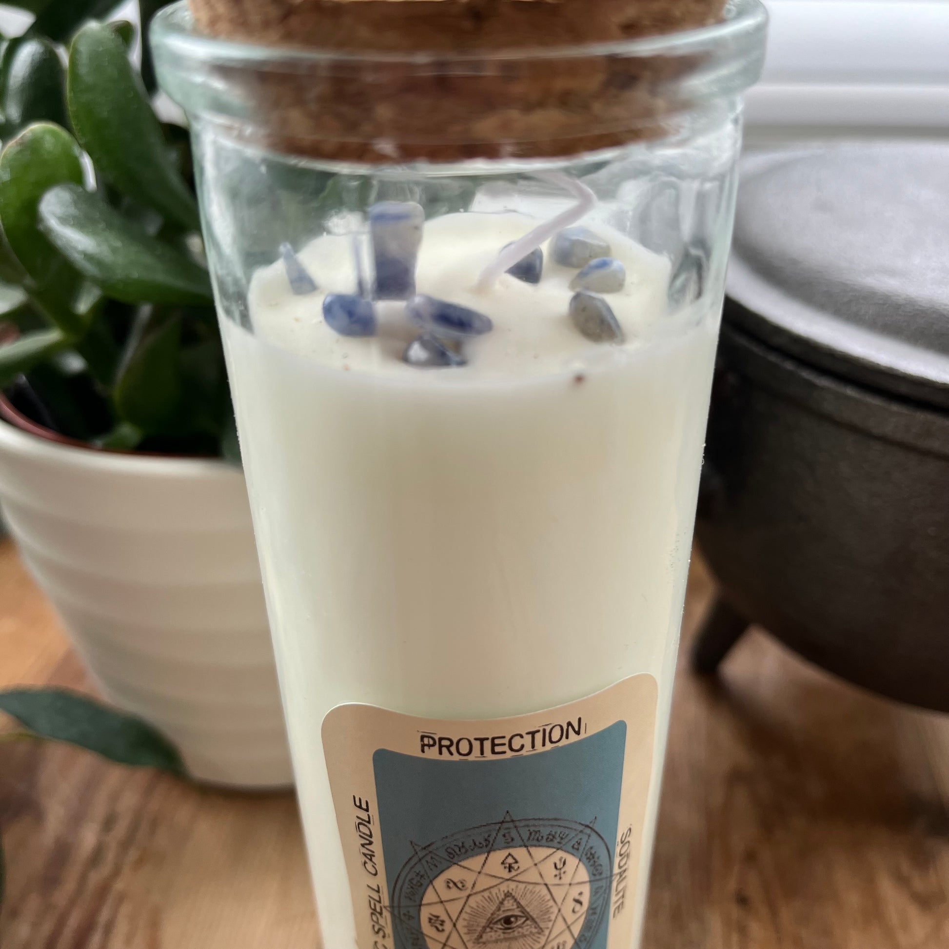 Tall glass spell candle with cork lid. The label on the candle features an image of the all seeing eye and the words Protection Sodalite and Dragons Blood. The candle itself is white with Sodalite gemstones embedded within it.