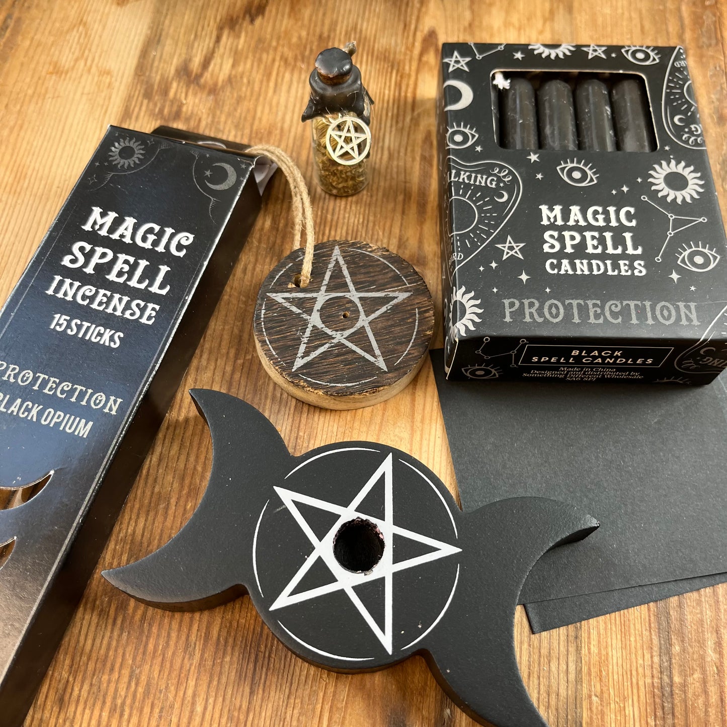 'Protection Spell Kit' Contents displayed on a wooden table. Pack of black opium scented incense sticks for protection and wooden pentagram incense holder, Pack of black magic spell candles, wooden black candle holder, in triple moon and pentagram design. squares of black paper and a "protection" spell bottle containing herbs and sealed with black wax and a charm.