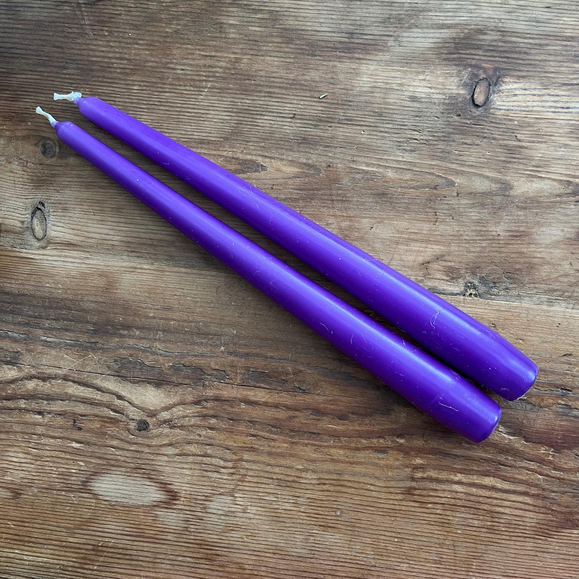 Two purple taper candles with white wicks, placed diagonally on a rustic brown wooden surface.