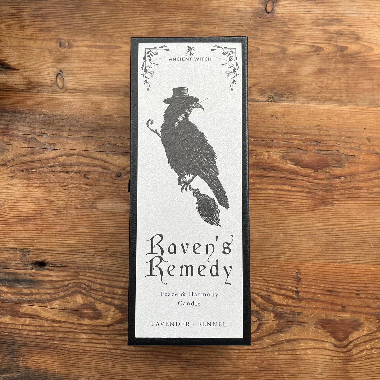 A framed candle box labeled 'Ancient Witch Raven's Remedy' with a black raven illustration, placed on a rustic wooden surface.