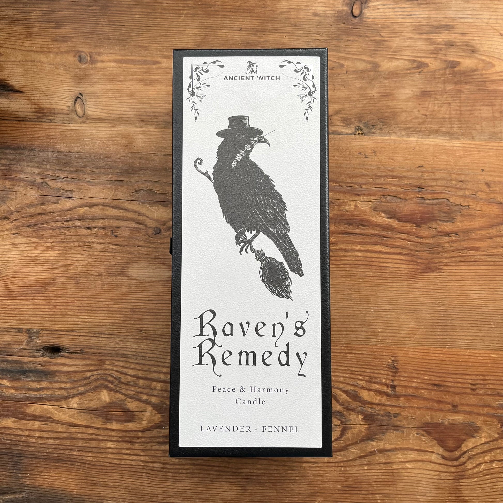 A framed candle box labeled 'Ancient Witch Raven's Remedy' with a black raven illustration, placed on a rustic wooden surface.