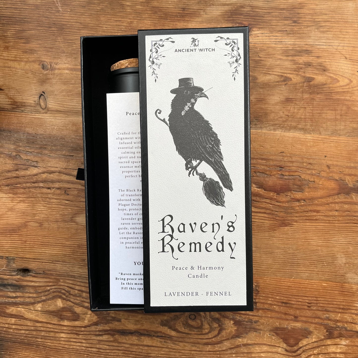 An open black presentation box containing a cylindrical candle jar and its front label, placed on a wooden surface. The label reads 'Ancient Witch' and 'Raven's Remedy' with a black raven illustration and text for 'Peace & Harmony Candle' and 'Lavender - Fennel'.
